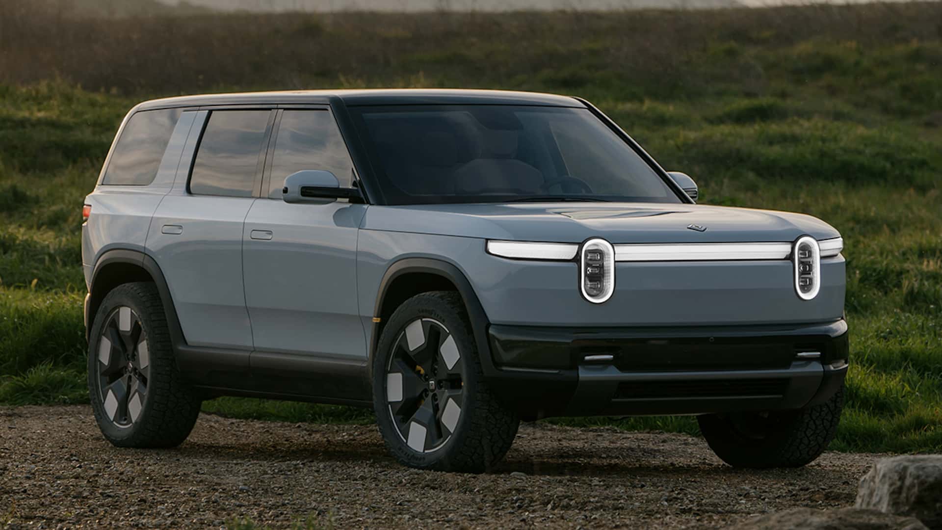 Vue extérieure du Rivian R2, un SUV électrique compact de couleur claire, garé dans un environnement naturel avec des arbres en arrière-plan.