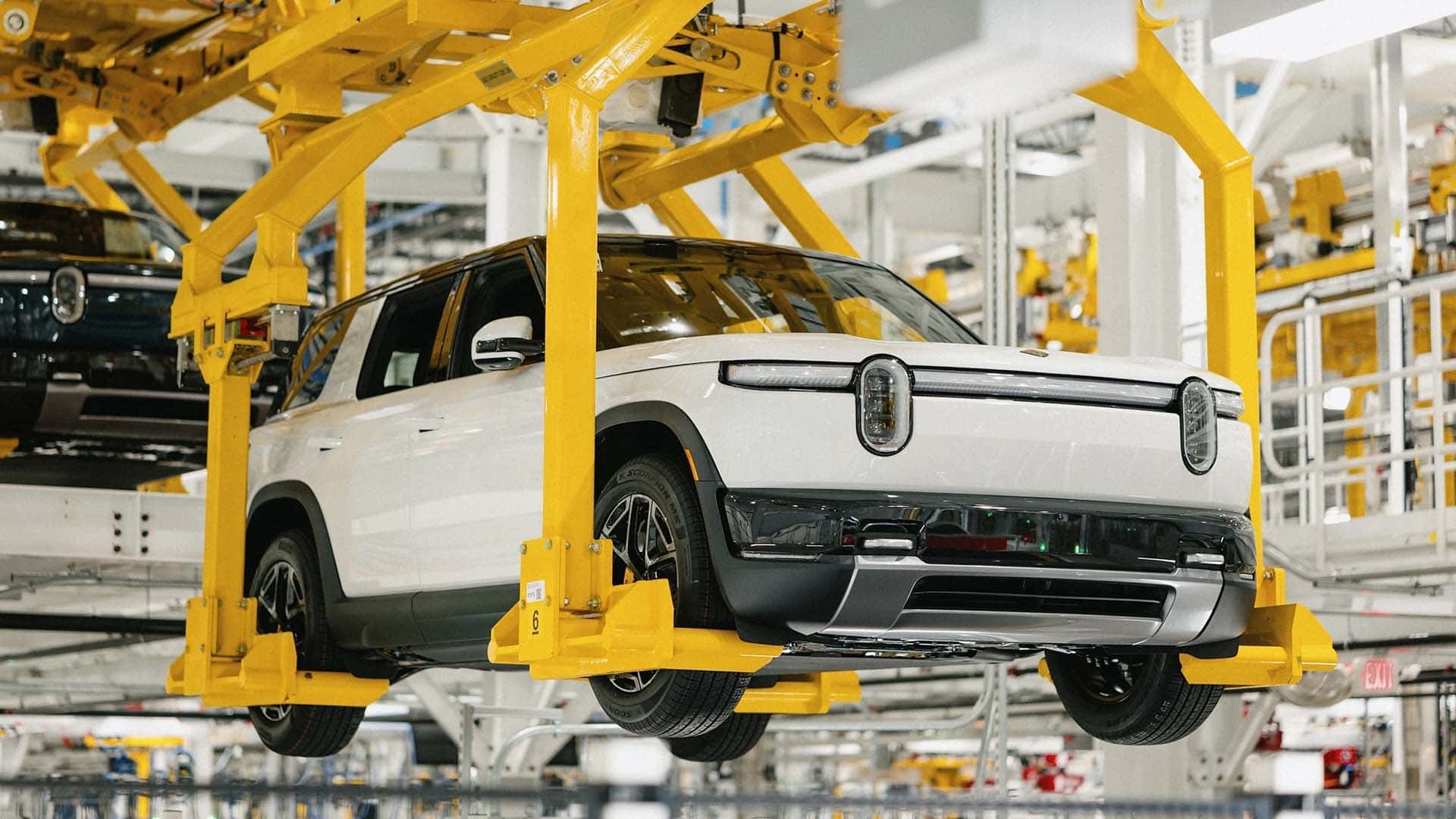 Image d'un Rivian R2 blanc sur une chaîne de production automobile, avec des robots et des ouvriers en arrière-plan.