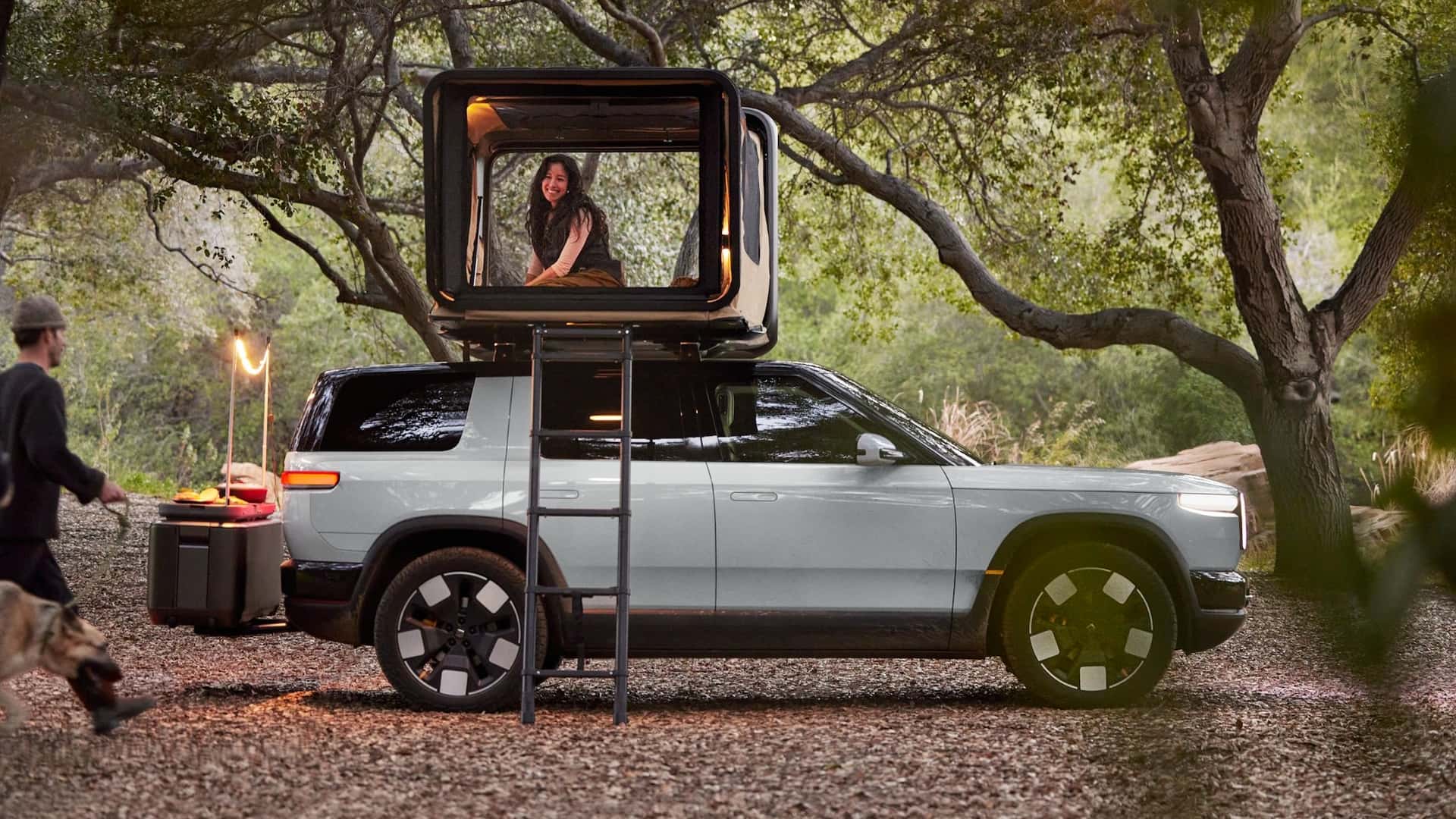 Un Rivian R2 vert foncé roulant sur un chemin de terre rocailleux, avec des arbres et des montagnes en arrière-plan.