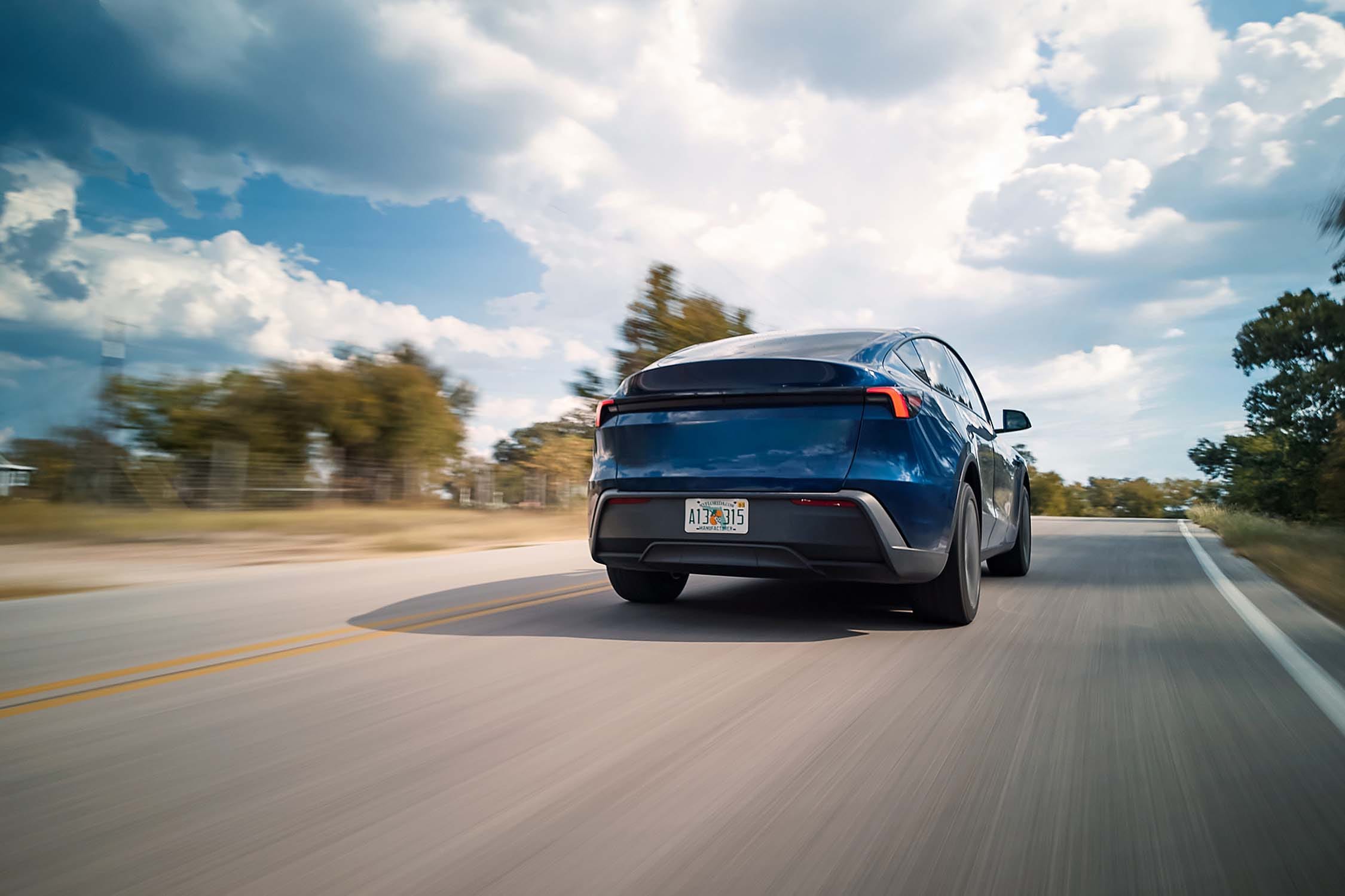 Un Tesla Model Y 2026 bleu roulant sur une route côtière au coucher du soleil.