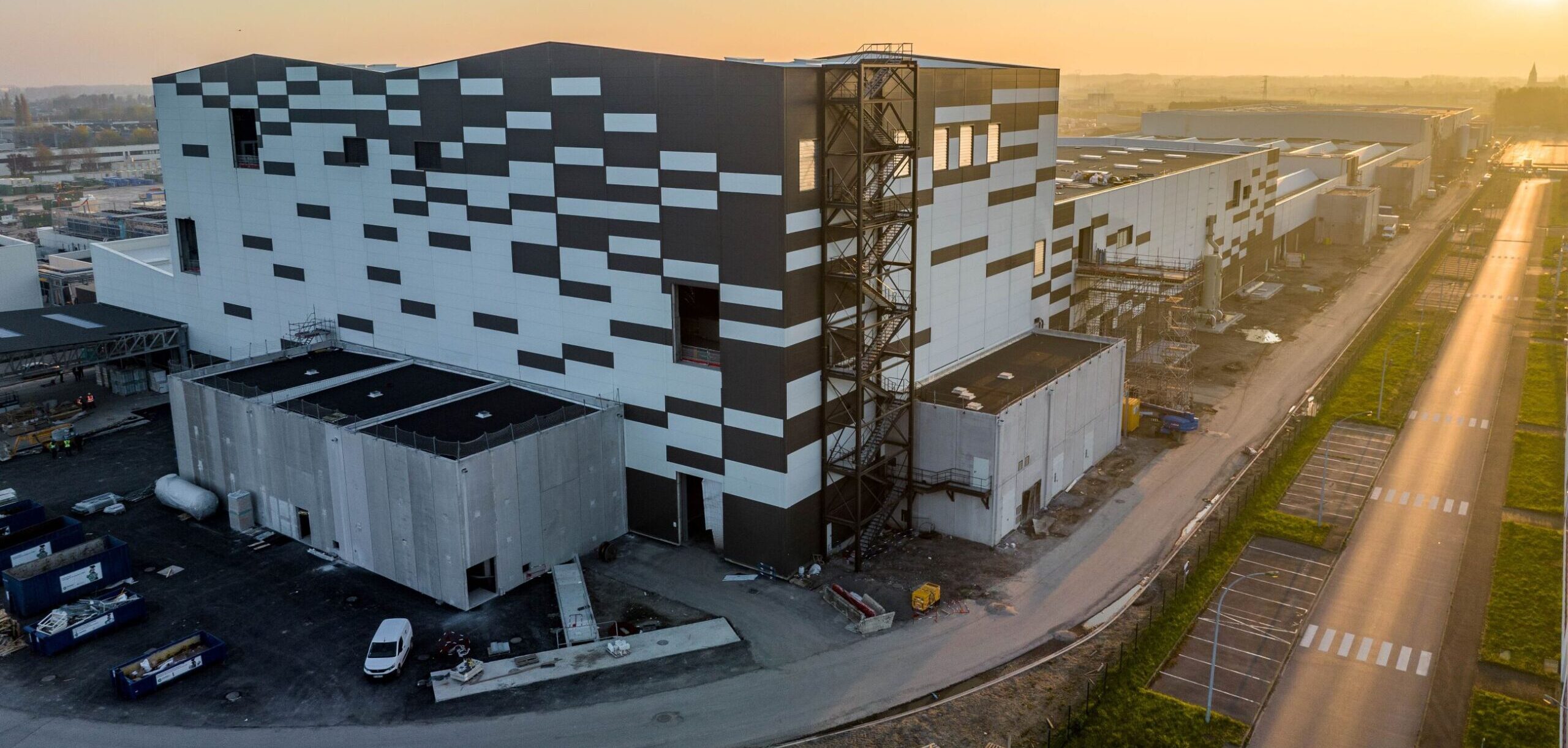 Vue extérieure d'une immense usine moderne avec des panneaux solaires sur le toit et des voitures électriques garées devant.