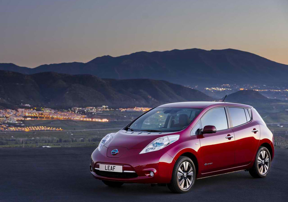 Une Nissan Leaf bleue foncée roule sur une route de ville, avec des bâtiments modernes en arrière-plan. La voiture est en mouvement, suggérant une conduite fluide et efficace.