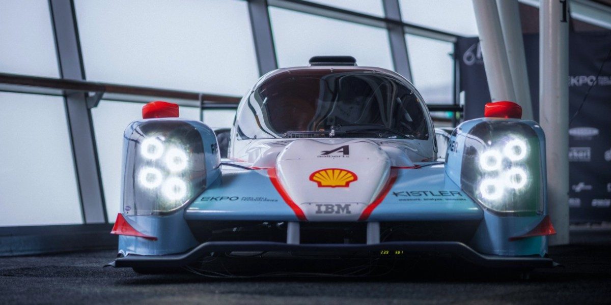 Photographie d'un arrêt au stand d'une voiture de course à hydrogène, avec des mécaniciens effectuant des tâches de ravitaillement et de maintenance.