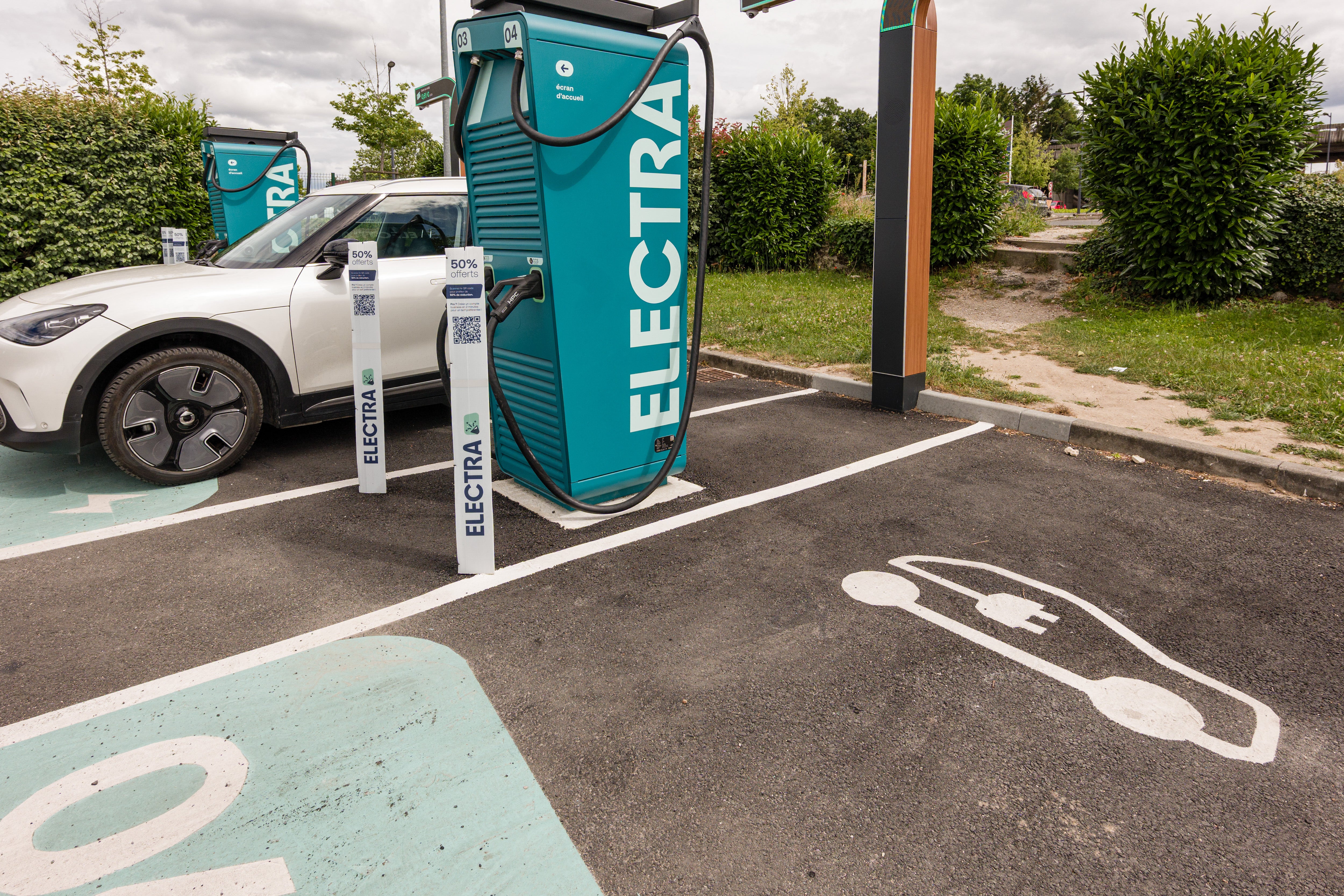 Une voiture électrique branchée à une borne de recharge rapide dans un paysage français ensoleillé. D'autres véhicules électriques sont visibles en arrière-plan, suggérant une station très fréquentée.