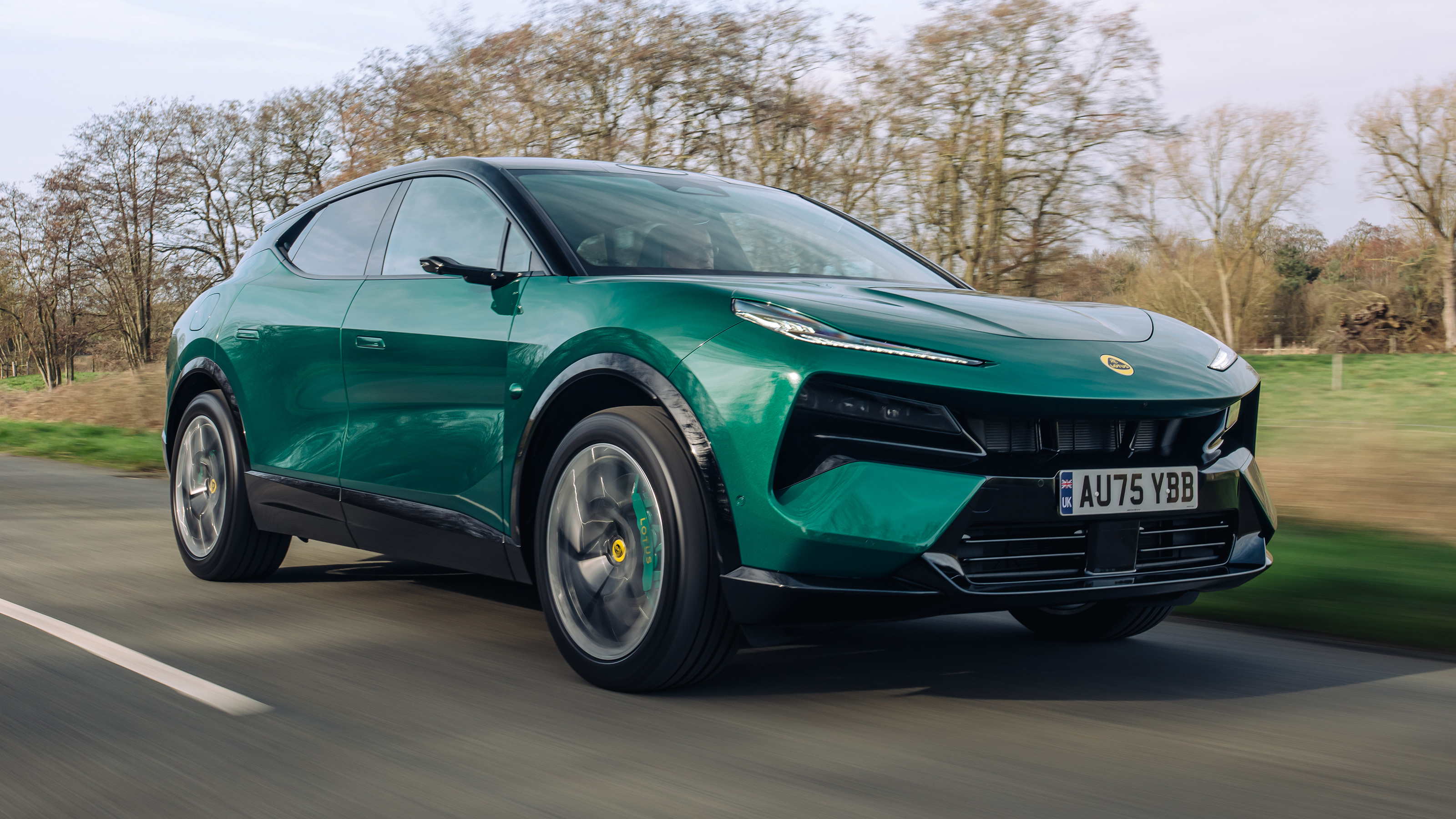 Vue latérale d'un SUV Lotus Eletre X gris foncé, en mouvement sur une route sinueuse, avec un arrière-plan flou de montagnes et de ciel bleu.