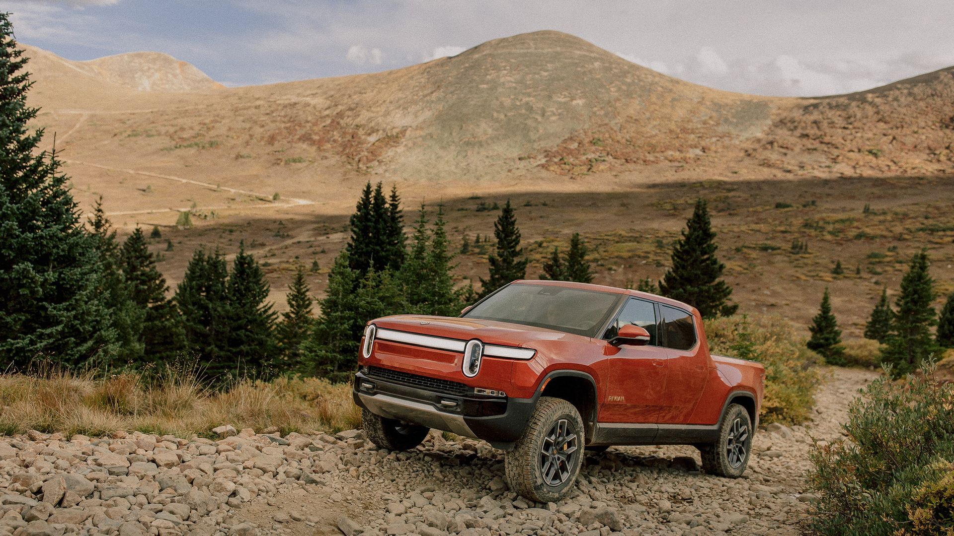 Un pick-up électrique Rivian R1T de couleur claire roulant sur une route sinueuse avec un paysage montagneux en arrière-plan.