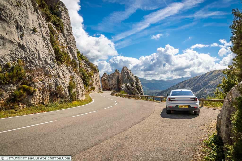 Une voiture électrique de couleur sombre roule sur une route de campagne sinueuse bordée d'arbres aux feuilles d'automne.