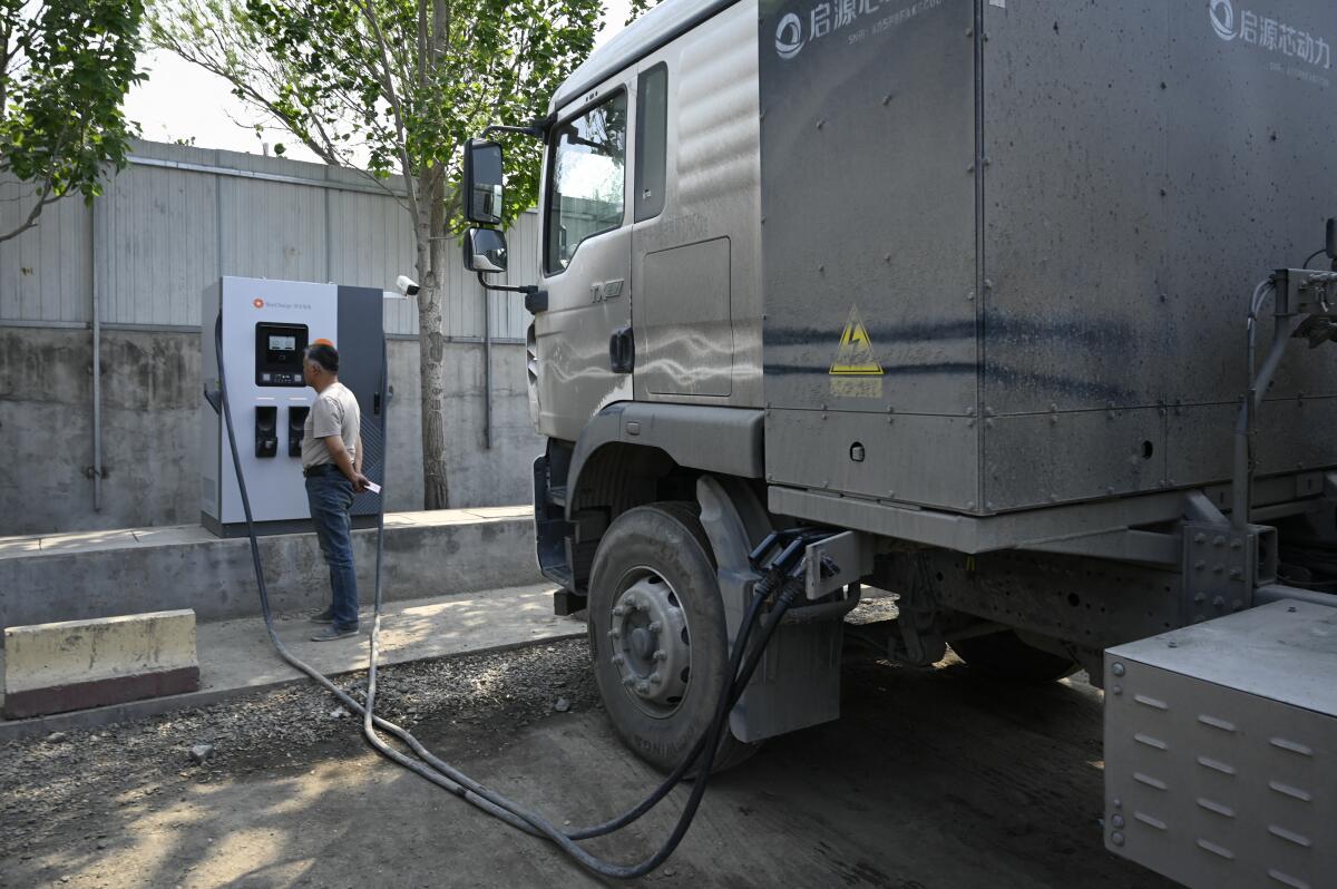 Plusieurs camions électriques chinois, allant des fourgonnettes aux poids lourds, sont connectés à des bornes de recharge dans une station moderne et bien éclairée.