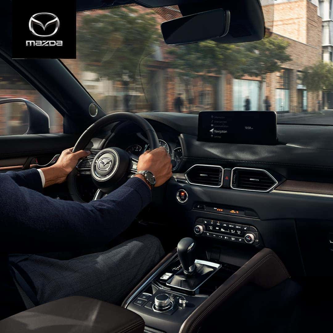 Vue intérieure du Mazda CX-5 2024 depuis le siège conducteur, montrant une main en train de gesticuler vers l'écran d'infodivertissement, suggérant une commande vocale.