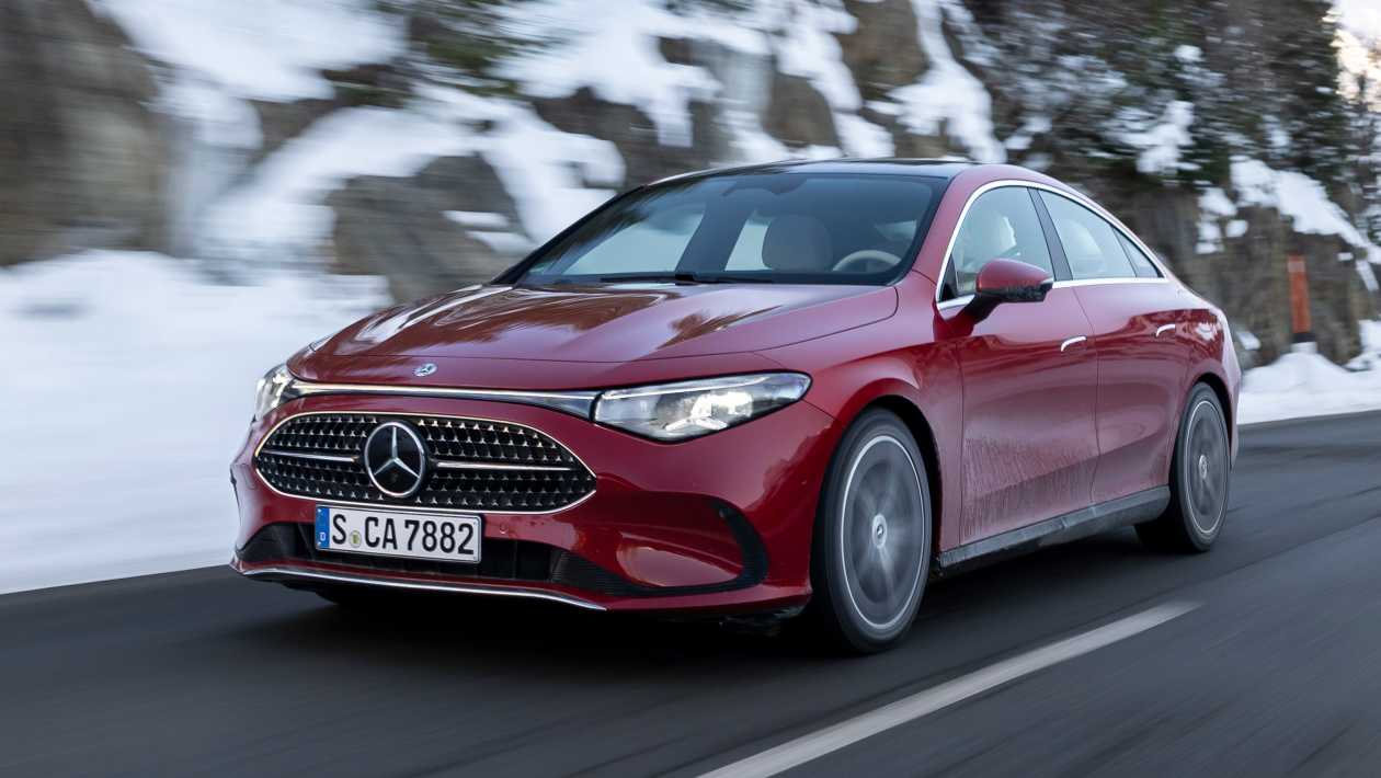 Une Mercedes CLA hybride 2025 de couleur sombre, en mouvement sur une route urbaine bordée de bâtiments modernes.