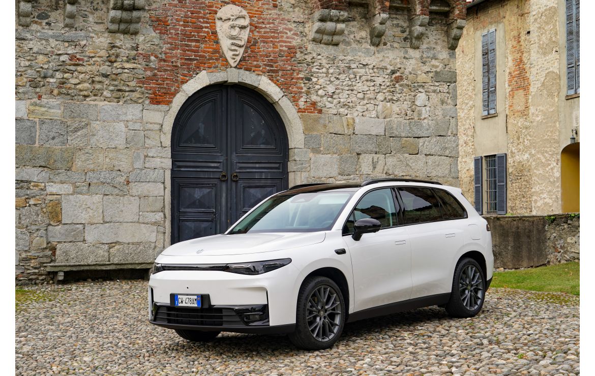 Une voiture électrique Leapmotor C10 blanche roule sur une route urbaine, avec des bâtiments modernes en arrière-plan.