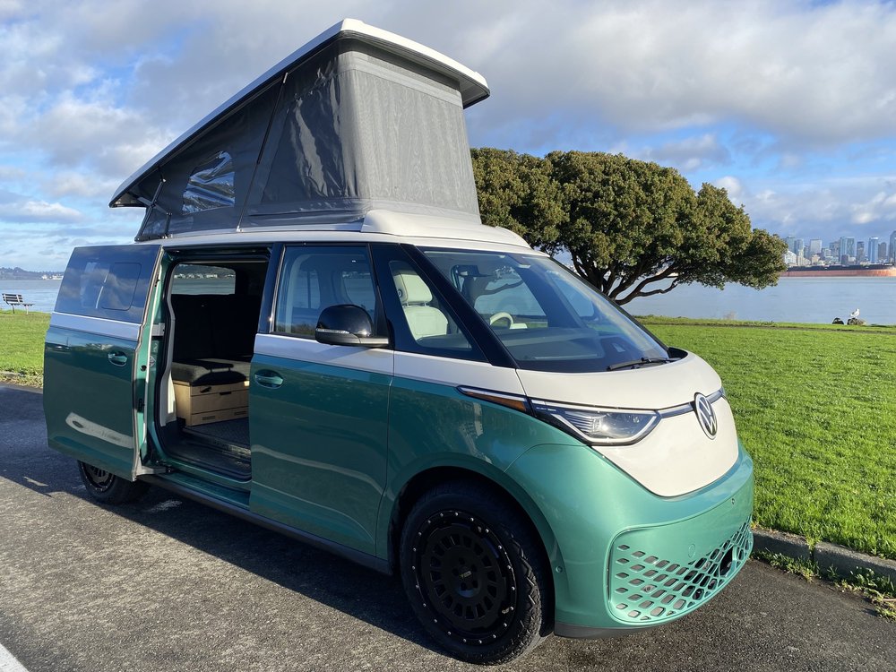 Un Volkswagen ID. Buzz personnalisé par Peace Vans, de couleur vive, garé dans un paysage naturel avec des montagnes en arrière-plan, mettant en valeur son design distinctif et ses équipements de camping.